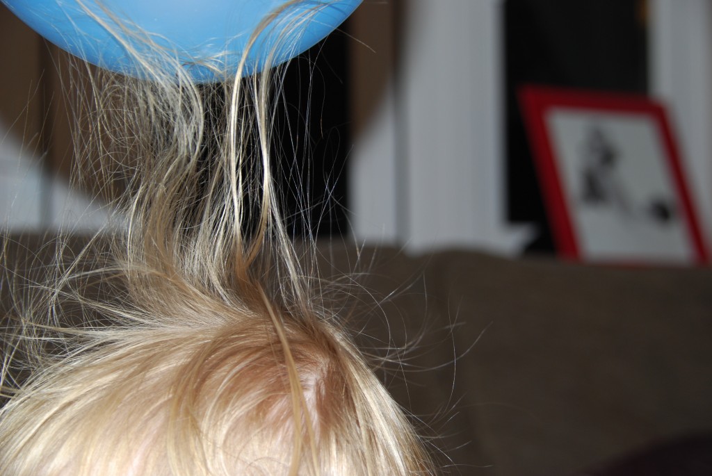 A child holding a balloon against their hair indoors, with strands of hair lifting toward the balloon from static electricity