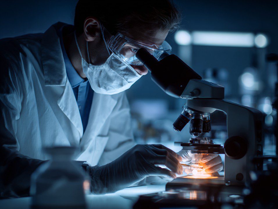 A laboratory photograph of a scientist working at a microscope workstation, with a computer monitor and lab bench tools visible nearby