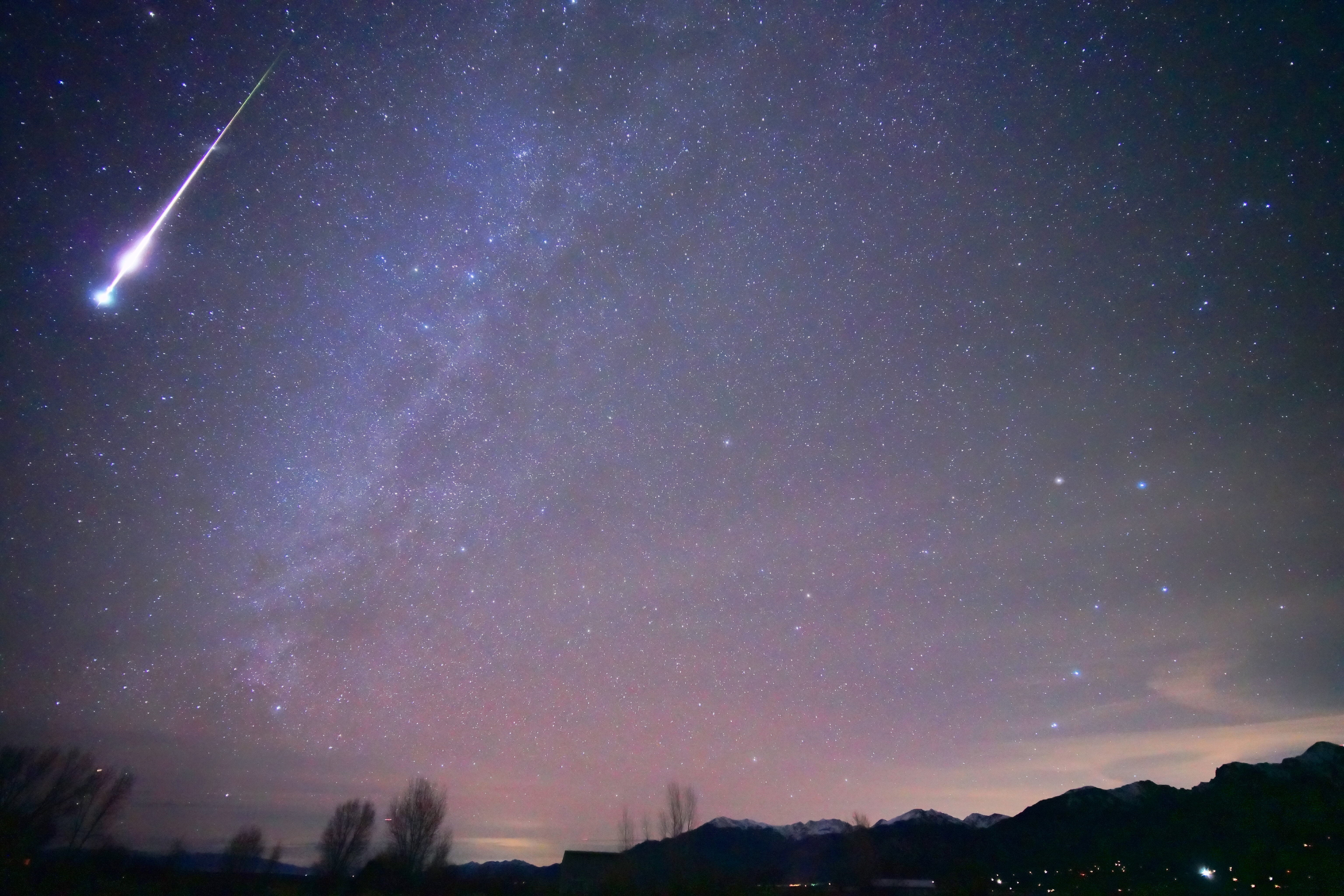 A night sky photograph showing a bright fireball meteor with a glowing trail above silhouetted trees