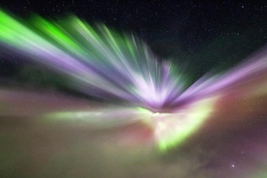 A real nighttime photograph of an aurora corona overhead in Iceland, with green rays radiating outward from a central point in the sky above a dark lava field