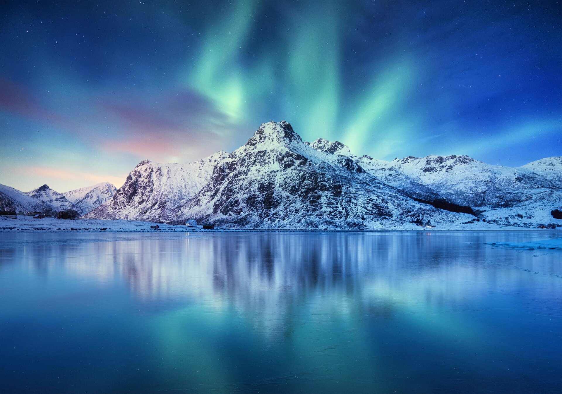 A real nighttime photograph of green aurora bands above the Lofoten Islands in Norway, with dark ocean water in the foreground and a small lit fishing village along the shore
