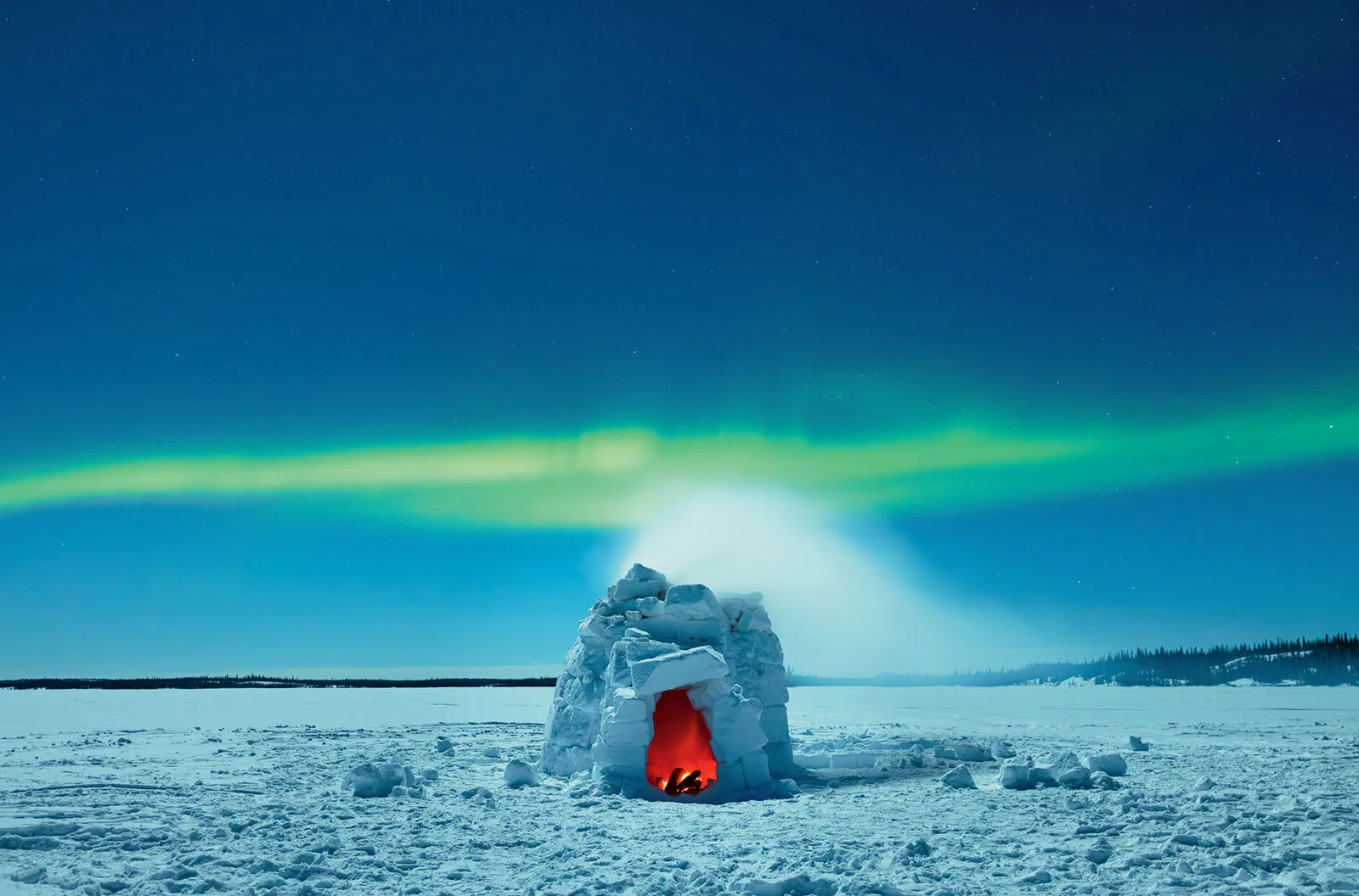 A real nighttime photograph of multicolored aurora with green and purple tones above a frozen lake near Yellowknife, Canada, with snow-covered shoreline and stars