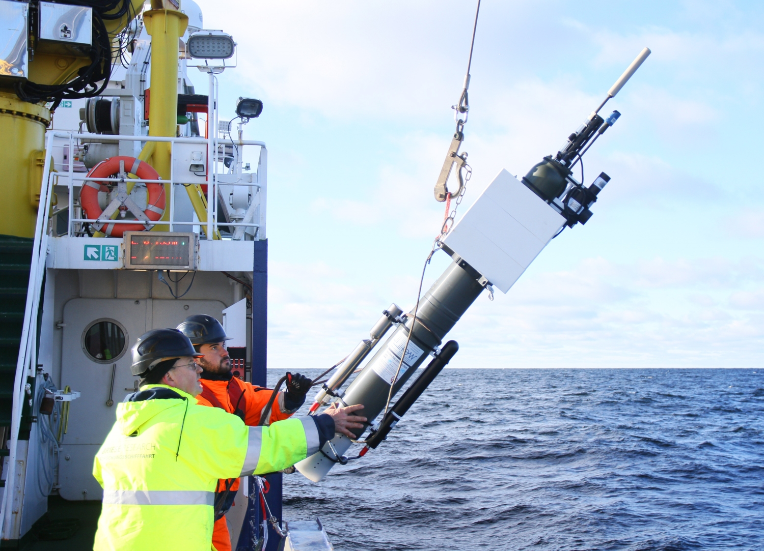 A researcher on the deck of an oceanographic vessel releasing a profiling float into calm blue water, with the horizon under a clear sky