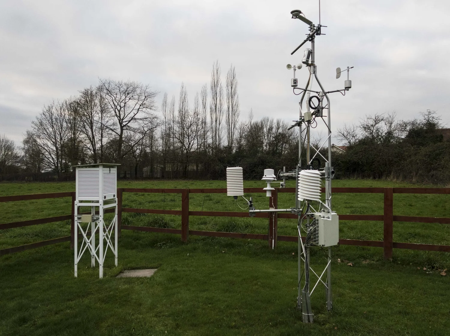 A weather station rain gauge and thermometer shelter in an open grassy field under a cloudy sky, with mountains faintly visible in the distance