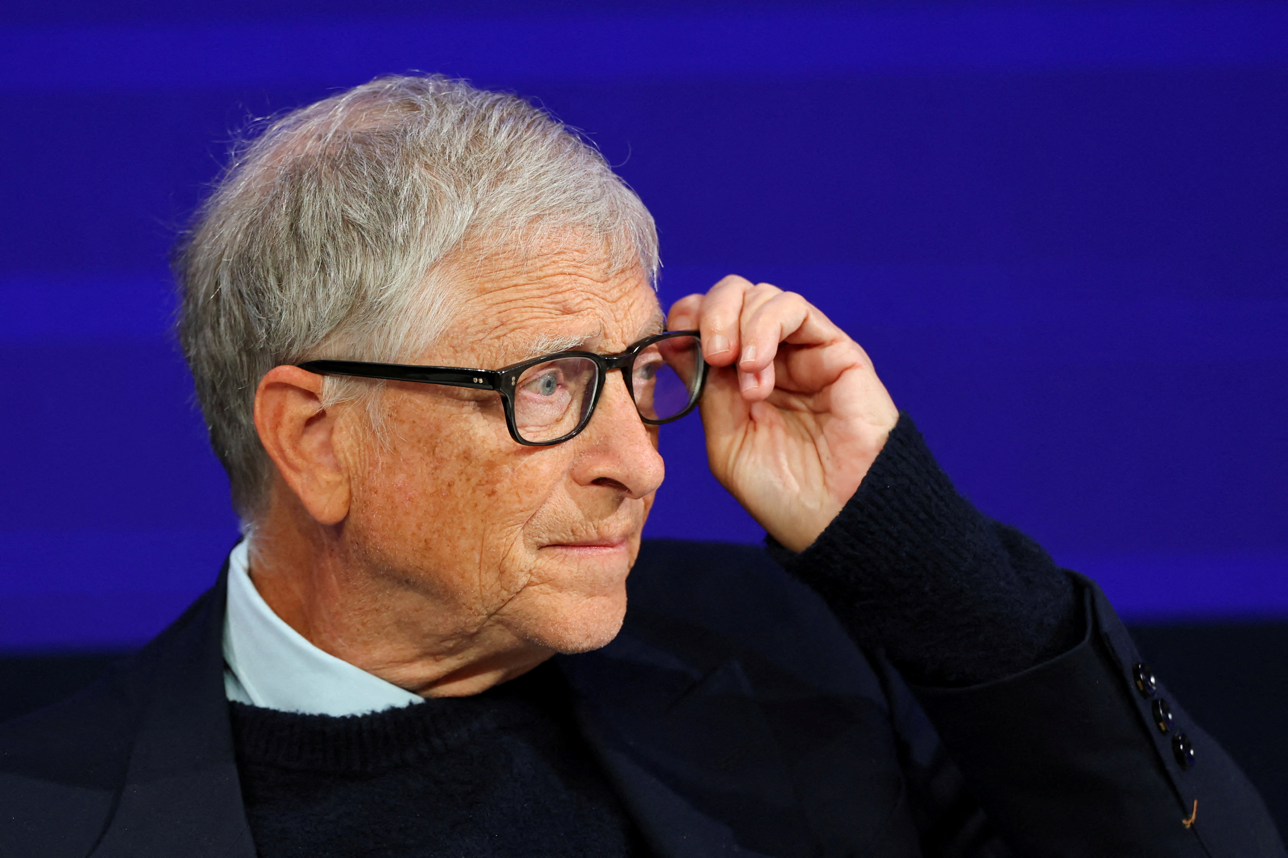 Bill Gates speaking on a stage during a public talk, standing under bright lights with a neutral backdrop, news photography style