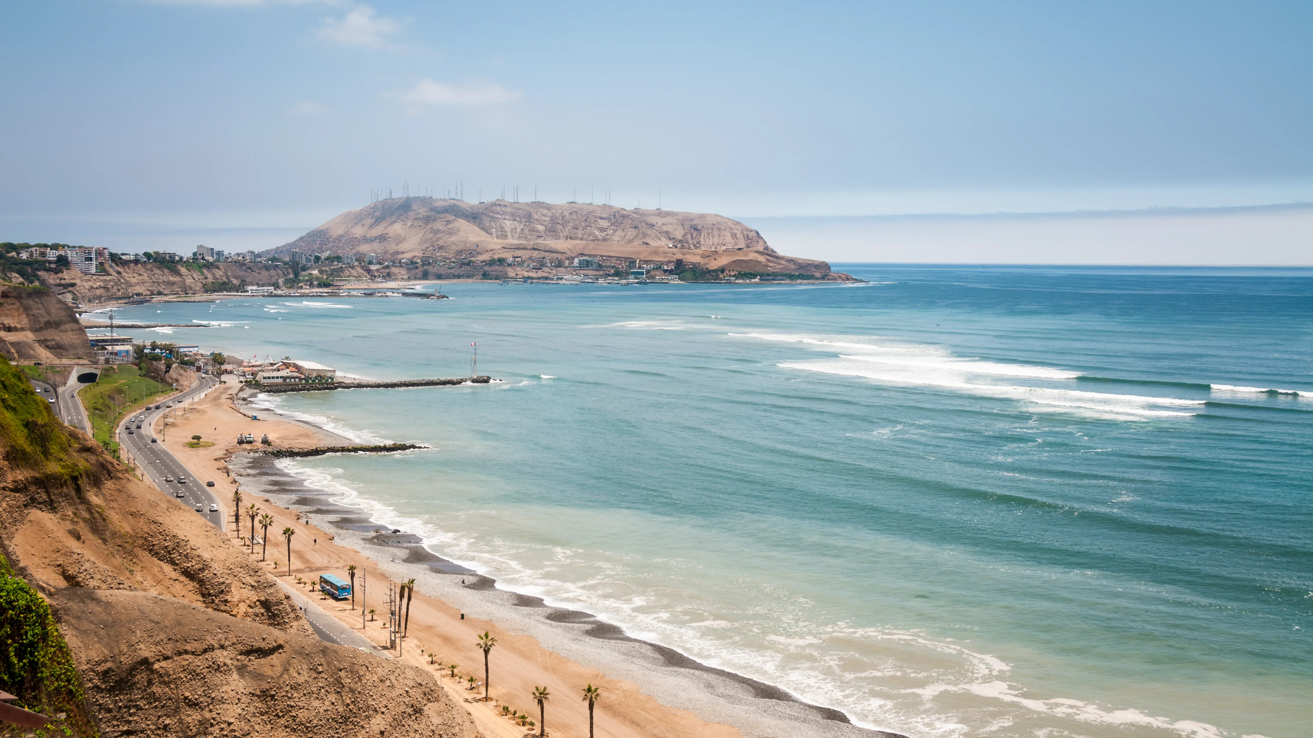 Waves breaking along a rocky stretch of the Peru coastline under a bright sky, with the open Pacific extending to the horizon
